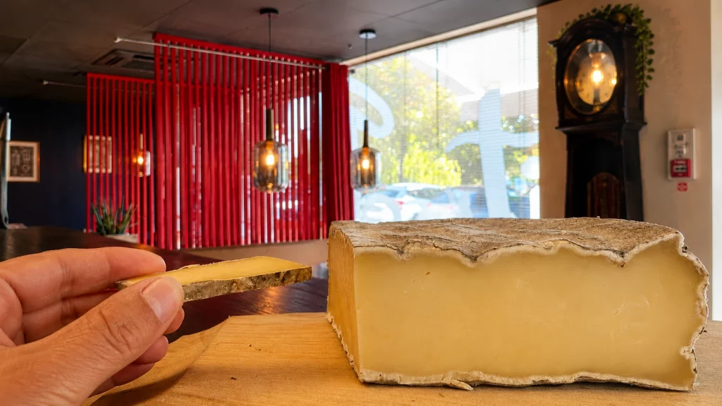 Morceau de croûte de Saint-Nectaire tenu entre les doigts devant un Saint-Nectaire au restaurant Let it Brie.